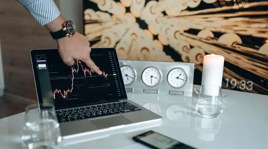 Hand pointing at market graph on laptop screen in modern office with clocks and candle.