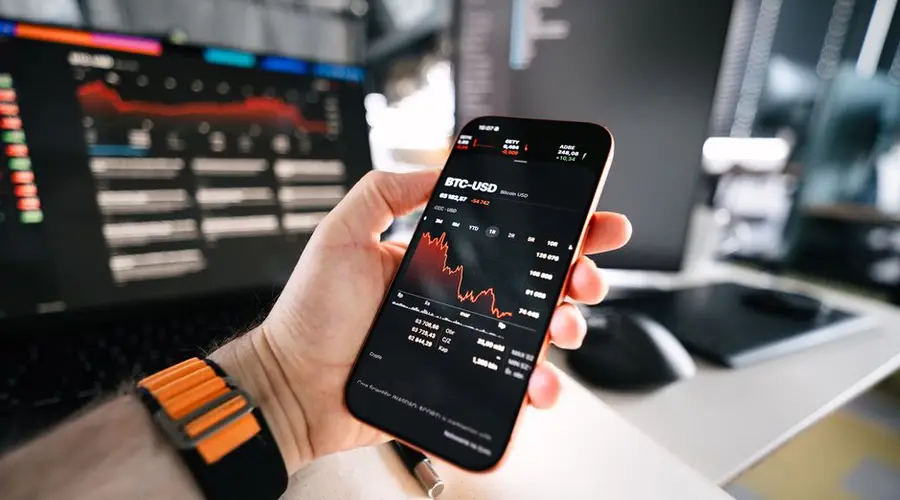 Hand holding smartphone displaying BTC-USD price chart with computer in background.