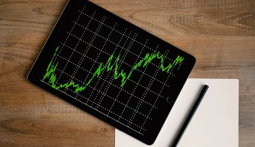 Flatlay of an iPad displaying stock market graph on a wooden desk with a pencil and paper.