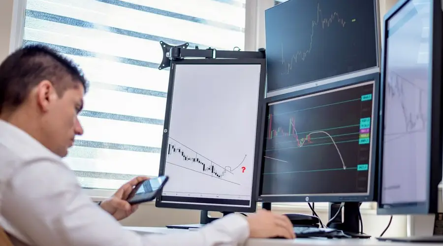 A stock trader analyzes financial data on multiple computer screens in an office setting.