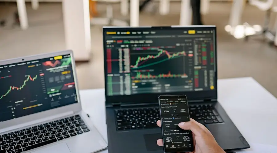 A professional tracks stock market trends using two laptops and a smartphone, showcasing digital finance tools.
