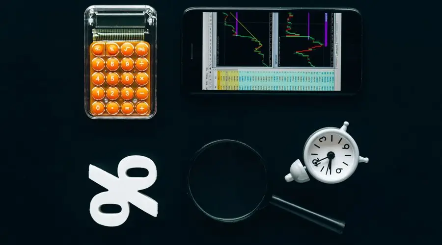 Top view of business tools including a calculator, smartphone with charts, magnifying glass, and clock on black background.