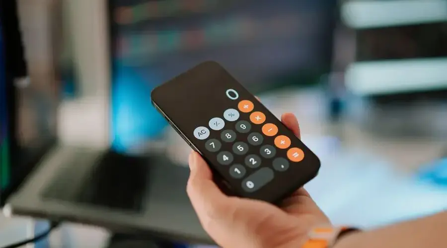 Close-up of hand holding smartphone calculator in office setting with financial data on screen.