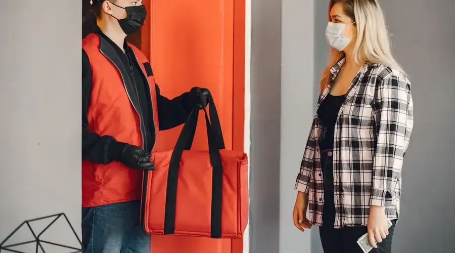 Young male courier in protective mask and gloves delivering order to young blond woman in checkered shirt and medical mask during safety food delivery at home