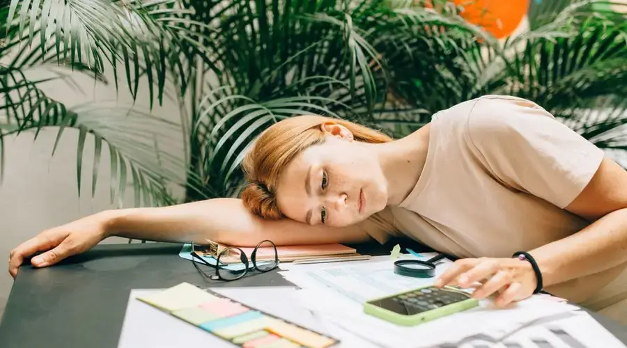 A tired woman calculating her taxes at a desk covered with papers and a smartphone.
