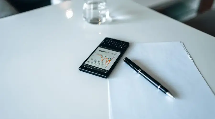 Smartphone displaying stock market graph on white desk with pen and paper.