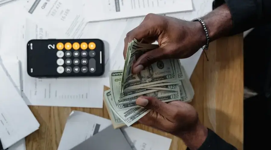 Person counting dollar bills over documents with a smartphone calculator on the desk.