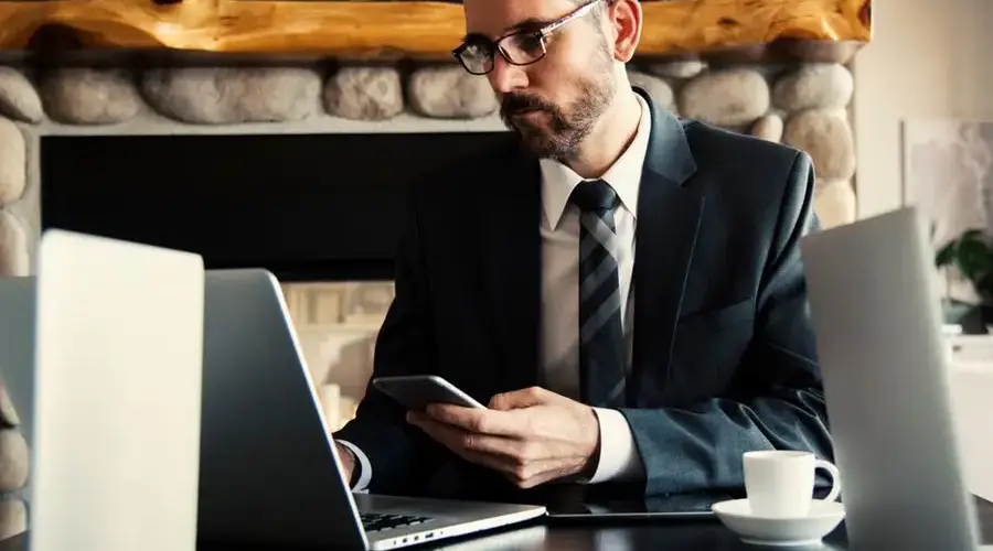 Businessman in formal attire working on a laptop and smartphone at an indoor office desk.