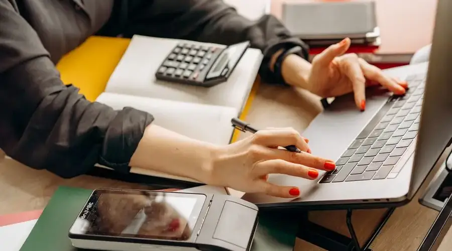 Close-up of a person using a laptop with a payment terminal and calculator on the desk.