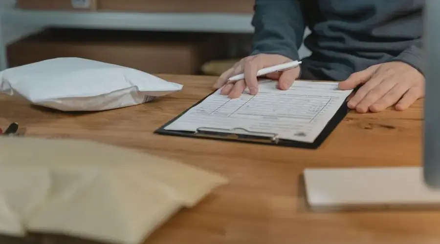 Close-up of a person filling out a form on a clipboard with packages nearby in an office setting.