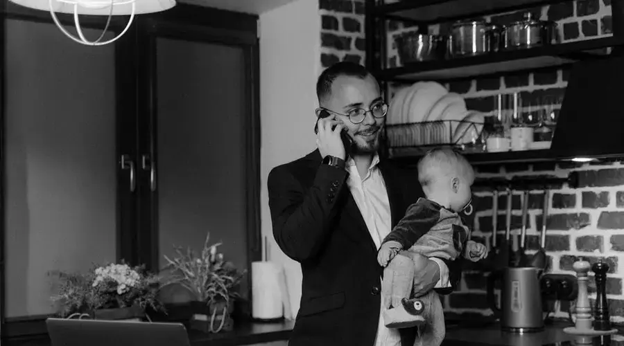 A father multitasks by holding his baby and using a phone in a modern kitchen.