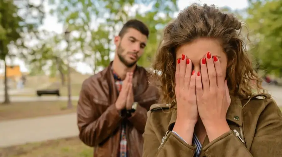 A young man apologizes while a woman covers her face, set in an outdoor park.