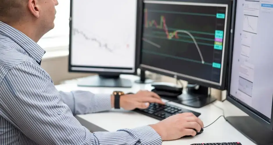 Businessman reviewing financial charts on multiple monitors in an office setting.