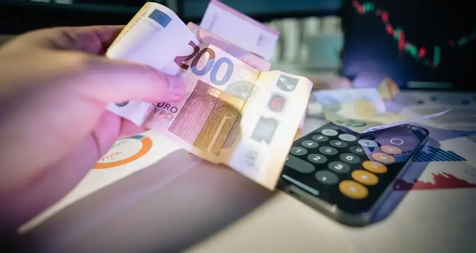 A hand holds euro notes with financial charts and a calculator, symbolizing investment and trading.