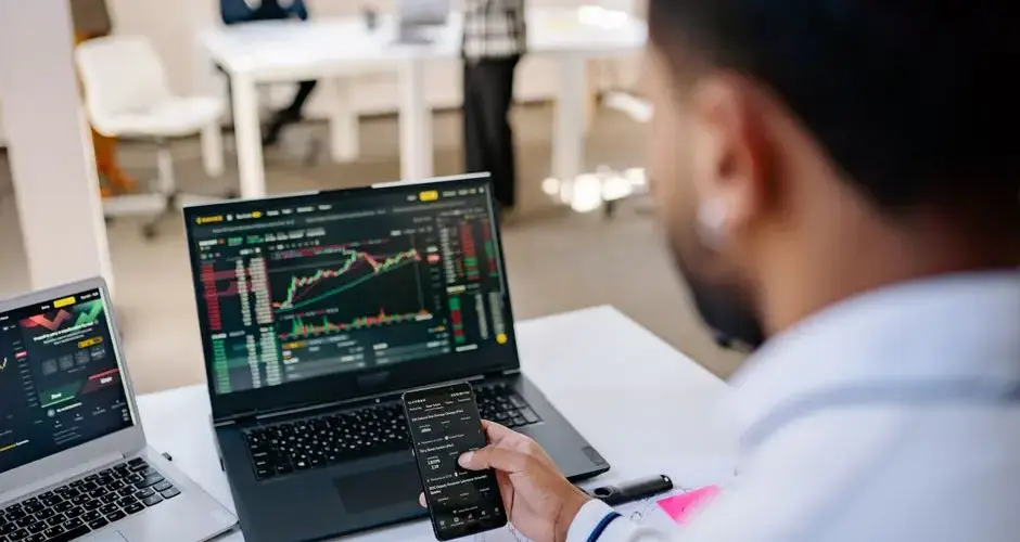 A businessman using smartphone and laptop to track stock market trends in a modern office setting.