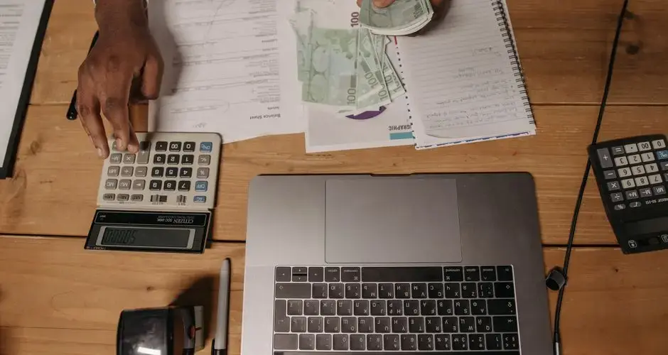 Close-up of a person counting cash with documents and a laptop in an office setting.
