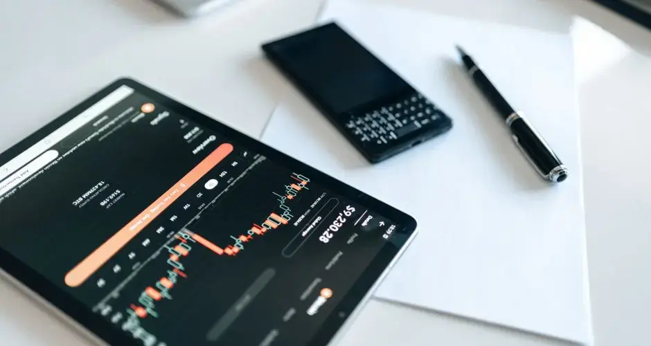 Close-up of a tablet displaying financial charts with a phone and pen on a white surface.