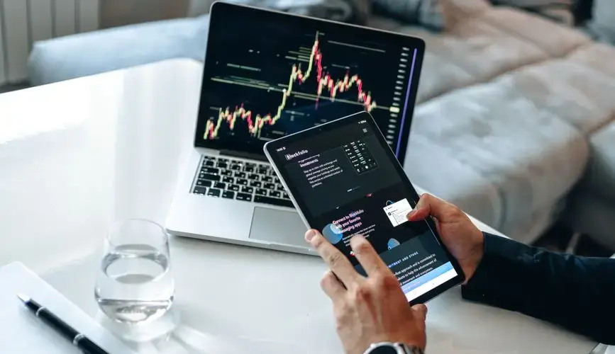 Person analyzing digital market trends on tablet with laptop showing financial charts.