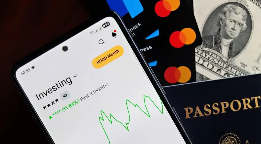 Smartphone displaying investing app, with credit cards, cash, and passport nearby, symbolizing finance