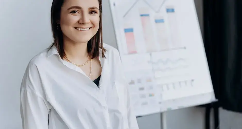 Cheerful woman in white shirt standing indoors with chart behind her, exuding professionalism.