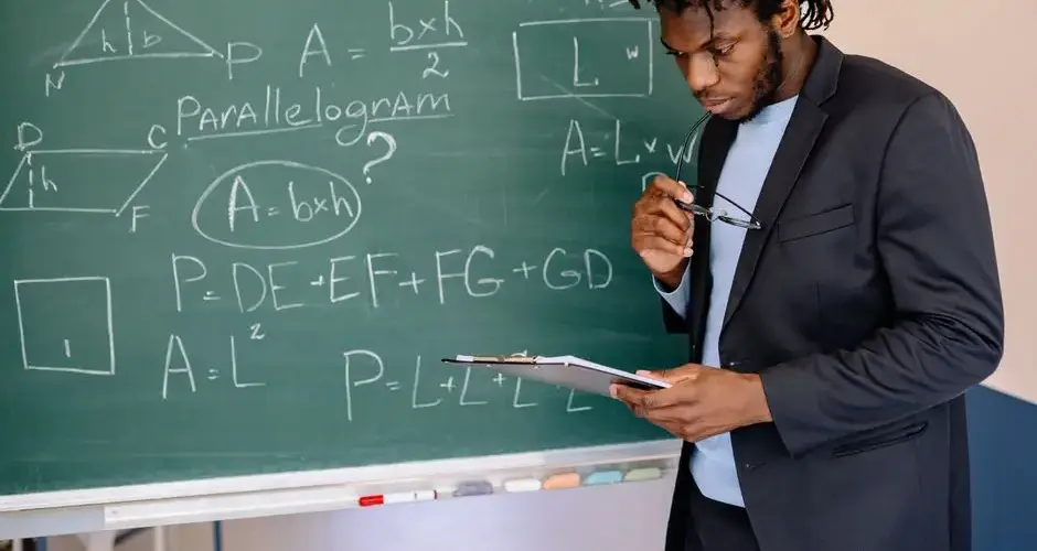Educator in suit jacket solving math equations on blackboard, holding clipboard.