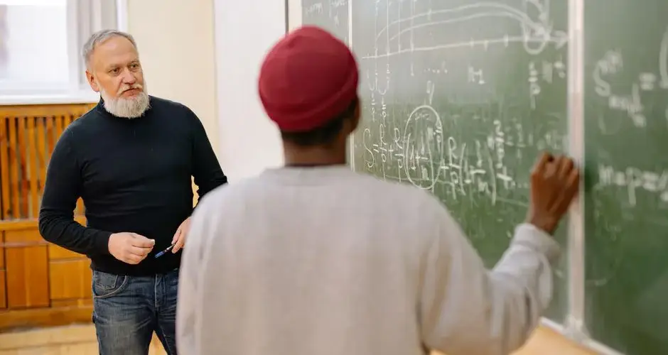 Educator assisting student during a math lesson at a blackboard in a classroom setting.