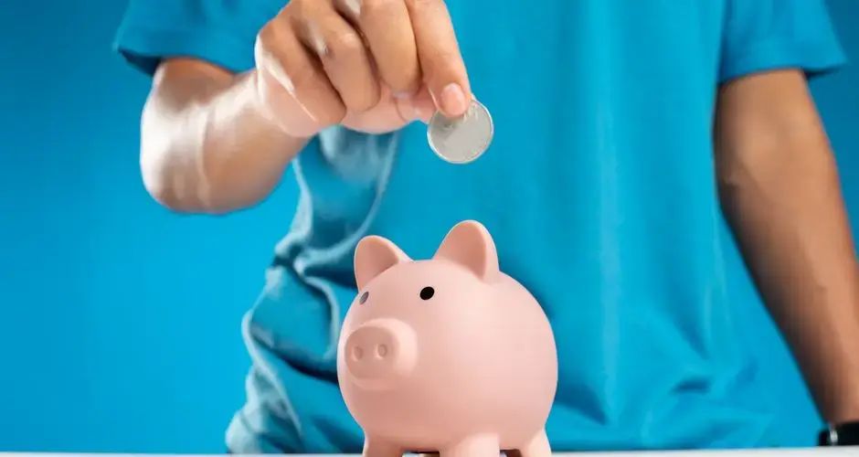 A close-up of an adult's hand dropping a coin into a piggy bank, symbolizing savings and investment.