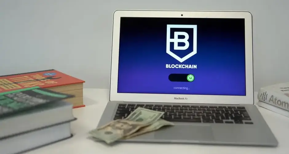 Laptop showing blockchain connection screen on desk with books and US dollars.