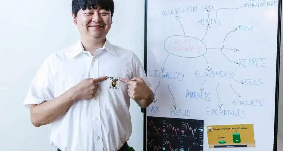Smiling businessman pointing at Bitcoin badge in front of whiteboard with cryptocurrency concepts.