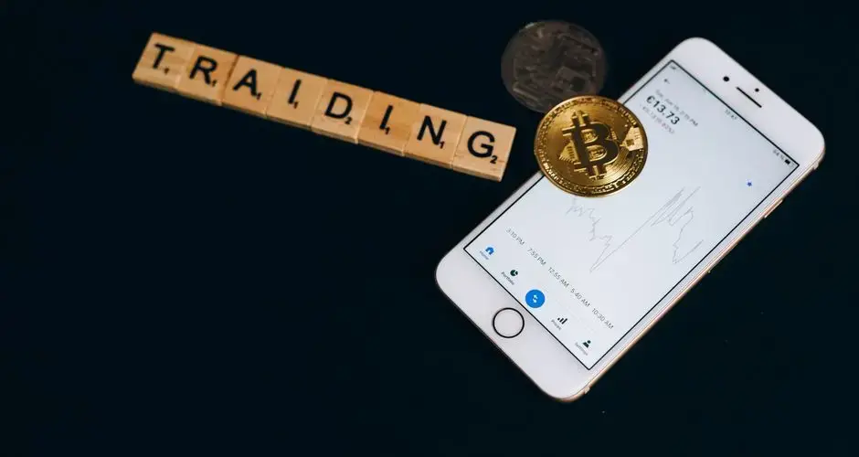 Cryptocurrency trading concept featuring Bitcoin, Scrabble tiles, and a smartphone on a black surface.