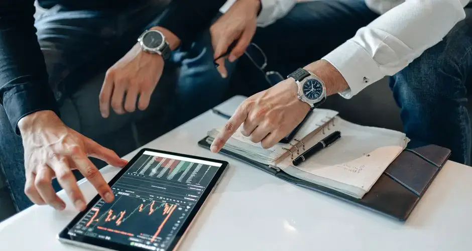 Close-up of professionals discussing stock charts on a tablet with notes and pens on a table.