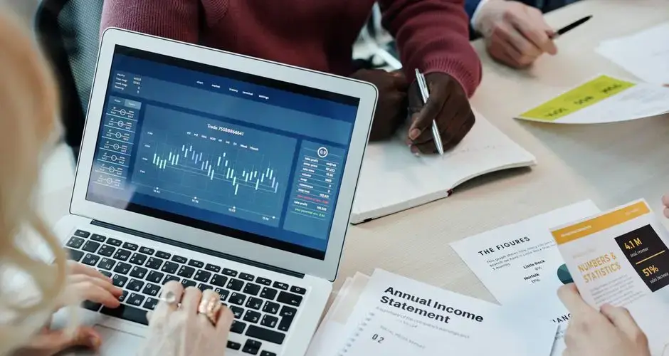 Business professionals analyzing financial data on a laptop during a meeting. Charts and reports visible.
