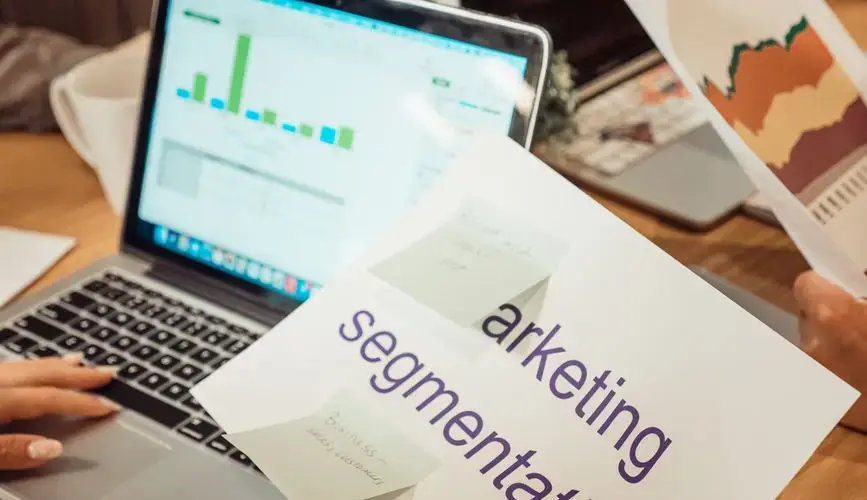 A modern workspace showing hands analyzing marketing data on a laptop and paper documents.