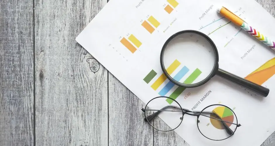 Overhead view of financial charts, a magnifying glass, ballpen, and eyeglasses on a wooden surface.