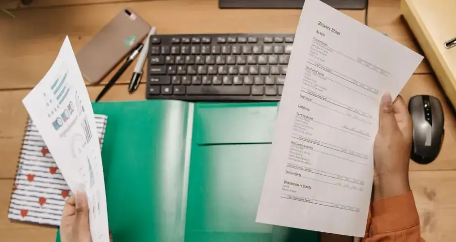 Hands holding and reviewing balance sheets over a desk with a keyboard and stationery.