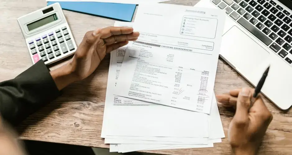 Hands holding financial documents with calculator and laptop on office desk, business analysis scene.