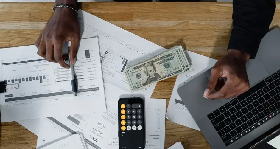 Hands calculate finances with papers, cash, and a laptop on a wooden desk.