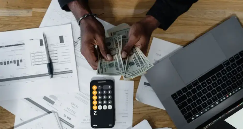 Person counting US dollars, using a calculator and laptop, with financial documents on a wooden desk.