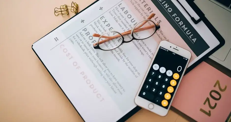 Overhead view of business tools including a phone calculator, pricing formula document, and eyeglasses on a desk.