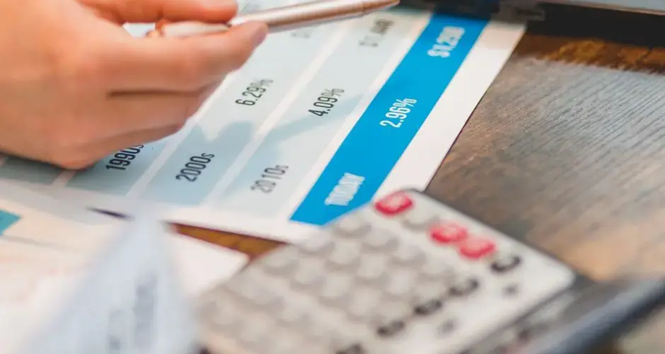 Close-up of a hand using a ballpen and calculator to analyze interest rates on a chart.