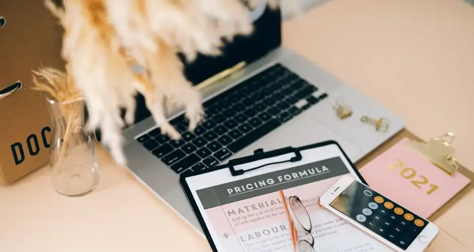 Organized office desk with pricing formula document, laptop, and smartphone calculator.