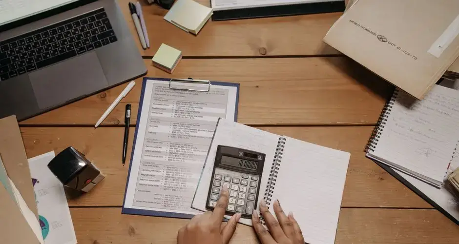 Hands working with calculator and documents on wooden desk in office setting.