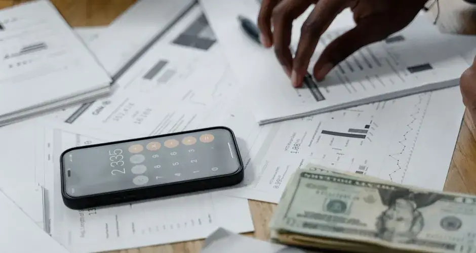 Close-up of hands calculating financial data with reports, smartphone, and cash on table.