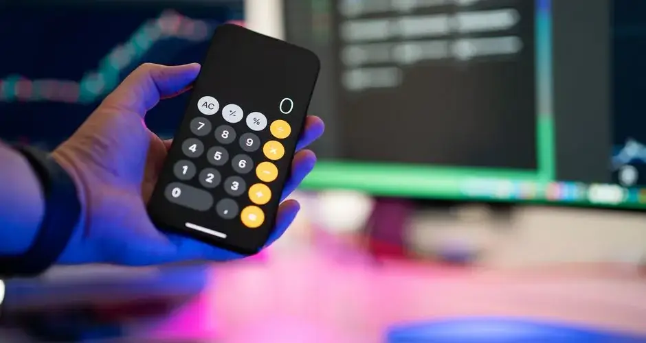 A person uses a smartphone calculator app in a modern office with computer screens displaying charts.