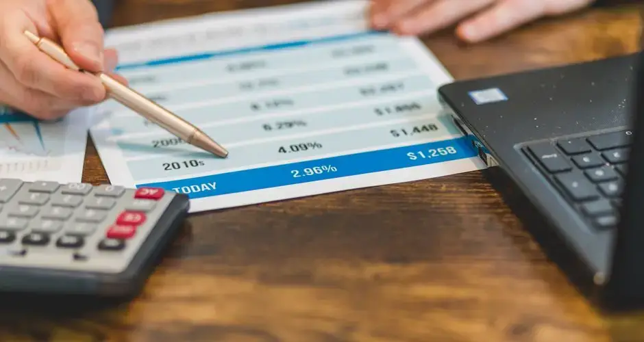 Close-up of a hand with pen analyzing financial rates on paper with a calculator and laptop nearby.