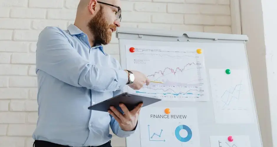 Man giving a business presentation with financial graphs on a whiteboard using a tablet for explanation.