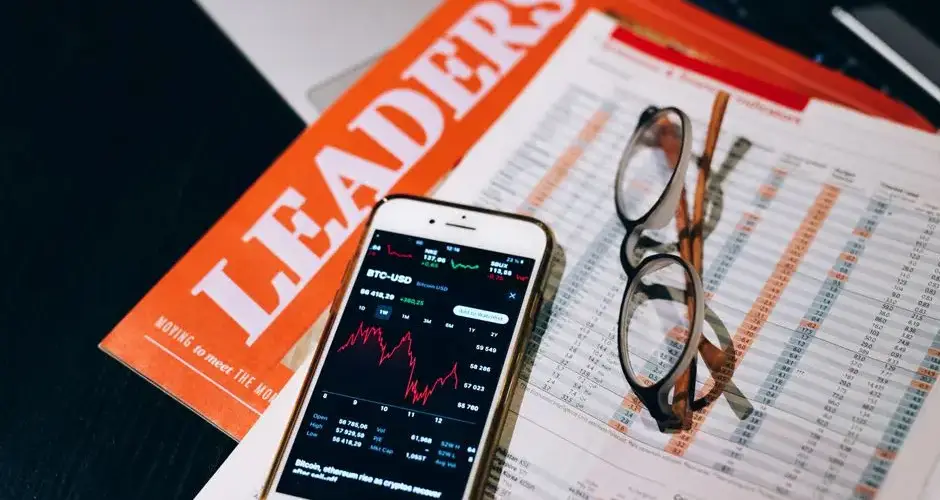 A close-up of a smartphone with cryptocurrency data next to financial reports and a magazine titled 'LEADERS'.