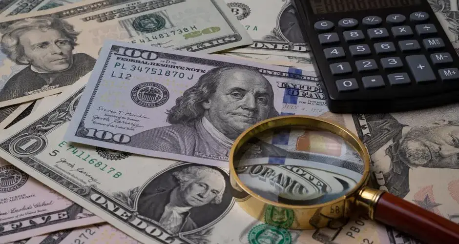 US currency bills with magnifying glass and calculator, symbolizing financial analysis.