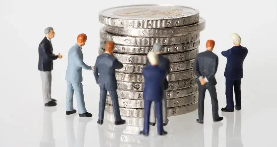 Miniature figurines of businessmen surround a stacked pile of coins on a clean white background.