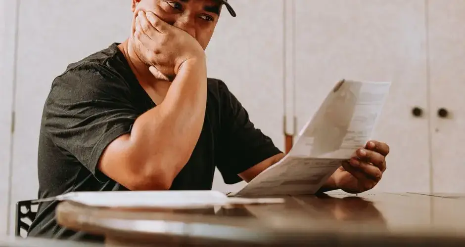 A man sits indoors, deeply focused on reviewing paperwork, expressing concern and contemplation.